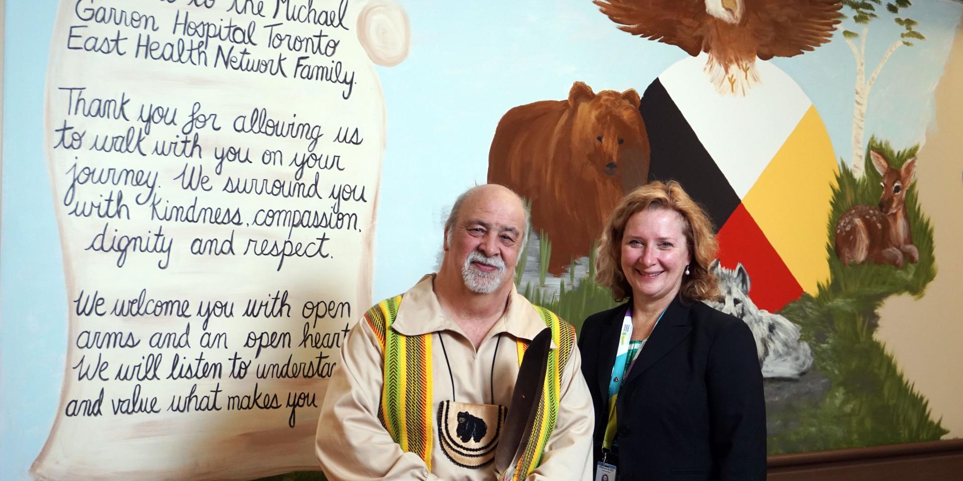 Ceo Sarah Downey and Elder Little Brown Bear in front of new medicine wheel mural at MGH