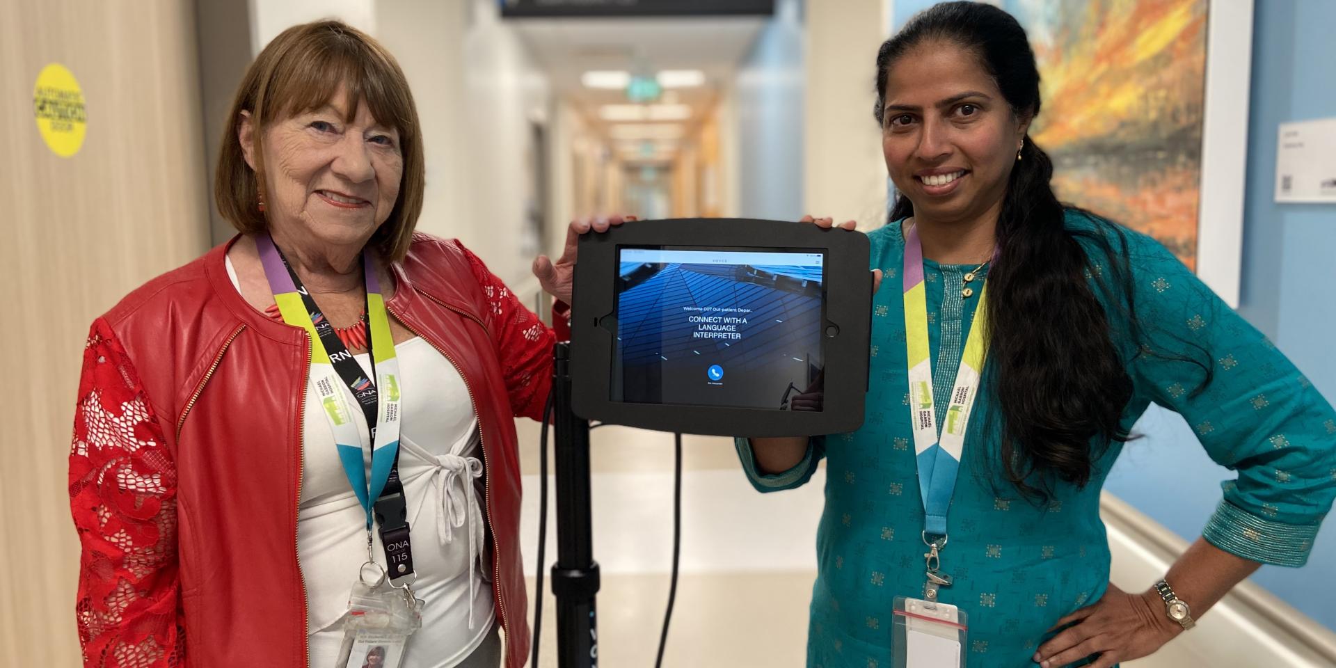 Two MGH staff members stand beside an iPad on a wheeled stand displaying the Voyce app