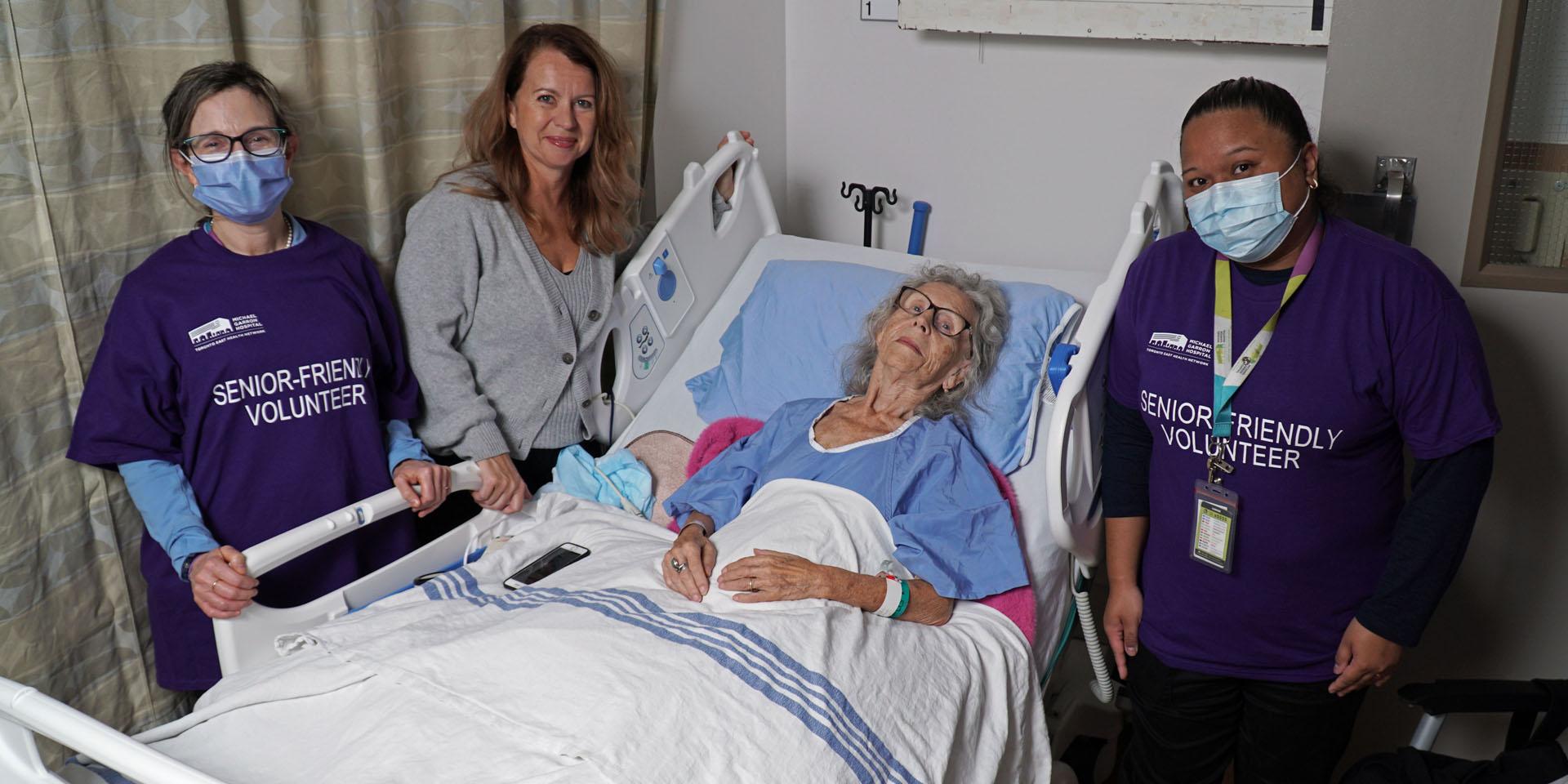 Two masked hospital volunteers standing next to an older patient in a hospital bed. The patient's daughter is smiling and also standing next to the bed.
