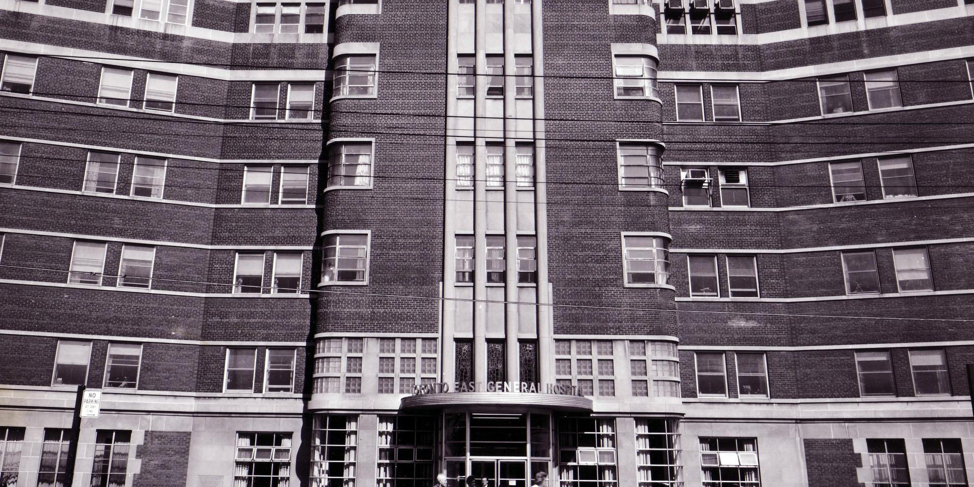 Black and white archival photo of Toronto East General Hospital entrance with vertical brick columns and curved wings extending on either side. Several people are walking near the entrance. 