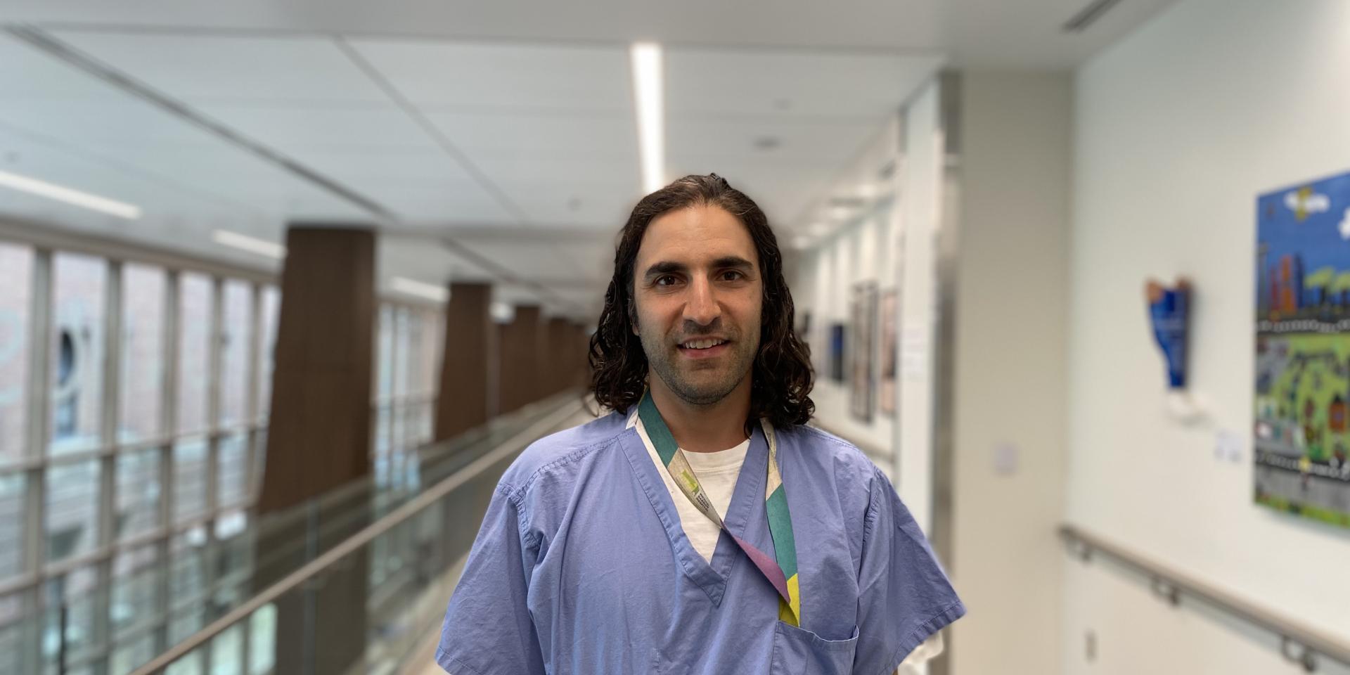 A man wearing hospital scrubs is standing in the hallway of a hospital looking directly at the camera.  
