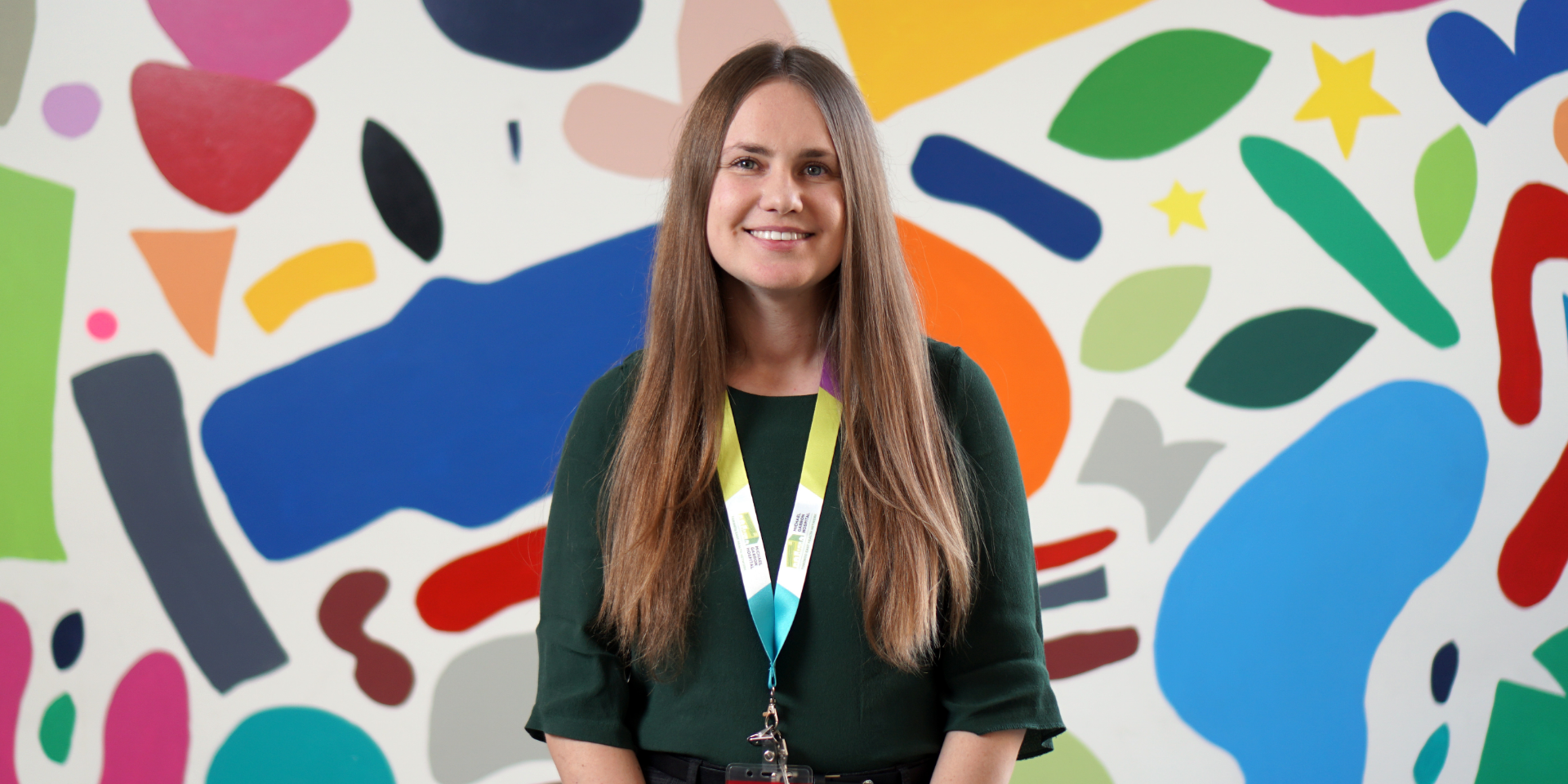 A woman smiling at the camera in professional attire in front of a colourful mural in a hospital.  