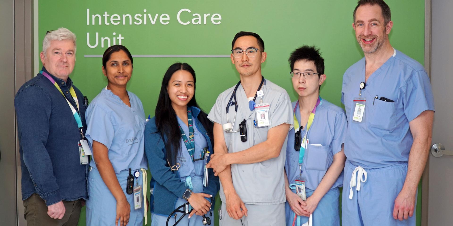 Six members of the ICU outreach team stand in front of a green wall with a sign saying "Intensive Care Unit." Five are wearing blue scrubs, all have hospital badges and are smiling at the camera.