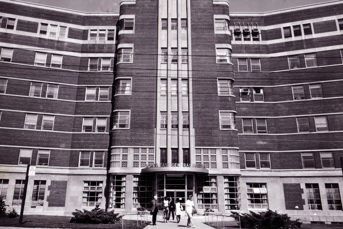 Black and white archival photo of Toronto East General Hospital entrance with vertical brick columns and curved wings extending on either side. Several people are walking near the entrance. 