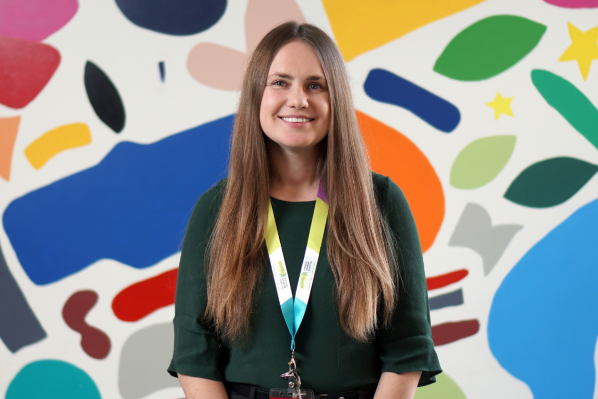 A woman smiling at the camera in professional attire in front of a colourful mural in a hospital.  