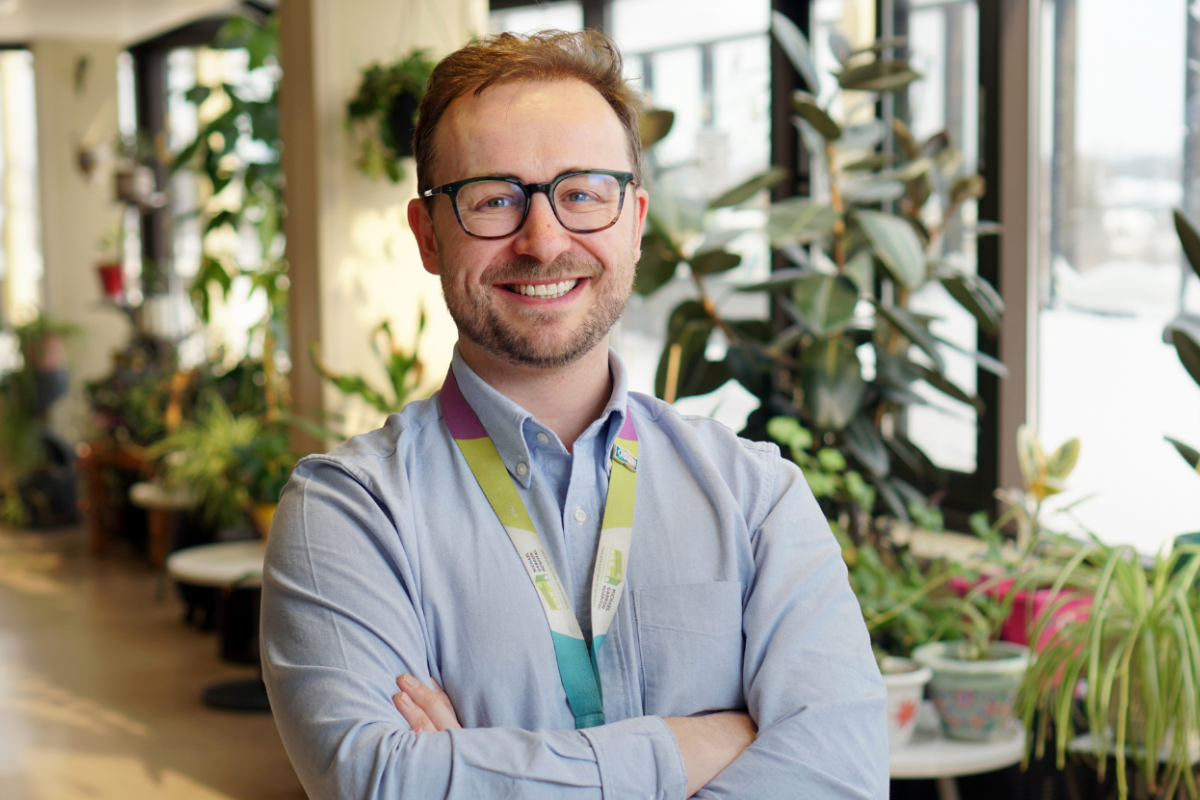 Man stands with arms crossed in room with plants. 