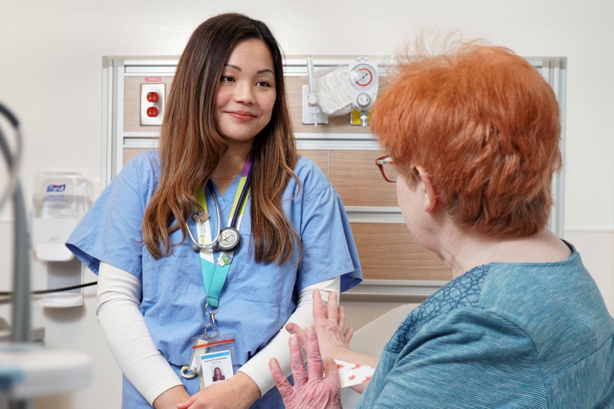 A female nurse in medical scrubs looks at an elderly woman in a hospital exam room. 