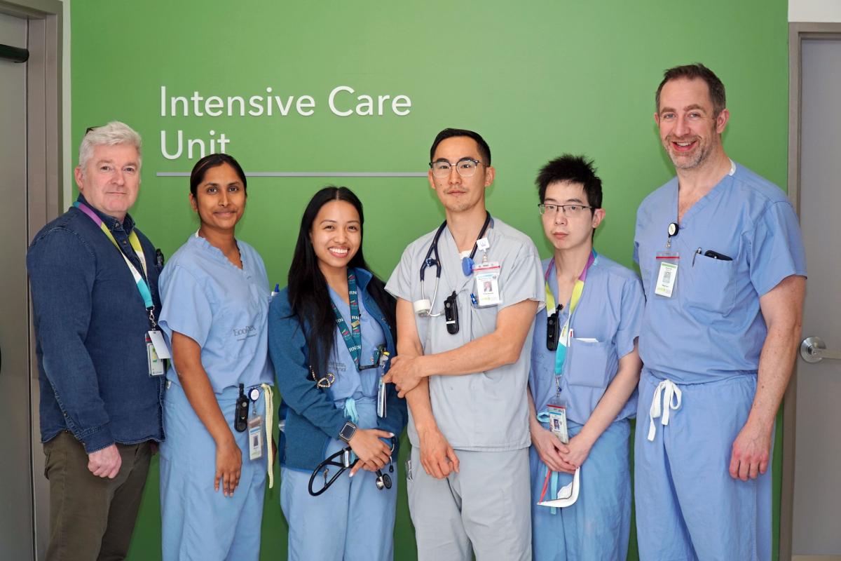 Six members of the ICU outreach team stand in front of a green wall with a sign saying "Intensive Care Unit." Five are wearing blue scrubs, all have hospital badges and are smiling at the camera.