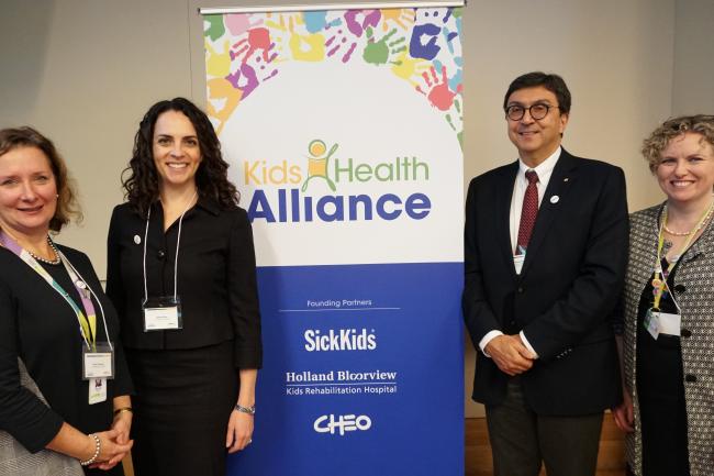 Four healthcare leaders stand in front of a stand-up banner that acknowledges the new partnership between Michael Garron Hospital and Kids Health Alliance 