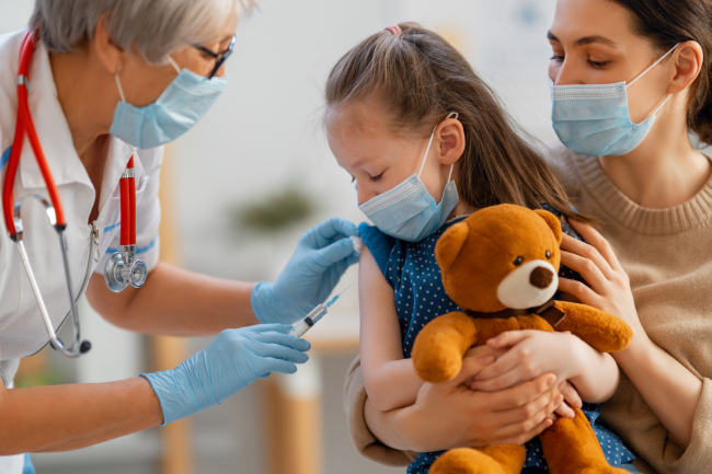 A child holds a stuffed bear while they are being vaccinated by a nurse.