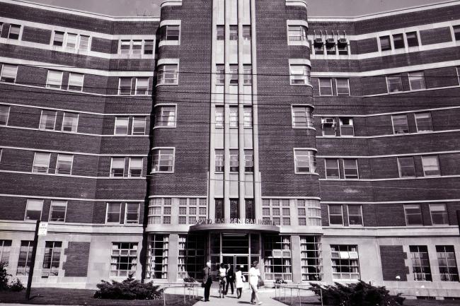 Black and white archival photo of Toronto East General Hospital entrance with vertical brick columns and curved wings extending on either side. Several people are walking near the entrance. 