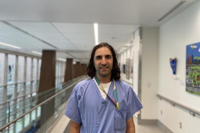 A man wearing hospital scrubs is standing in the hallway of a hospital looking directly at the camera.  