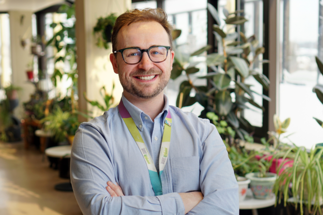 Man stands with arms crossed in room with plants. 