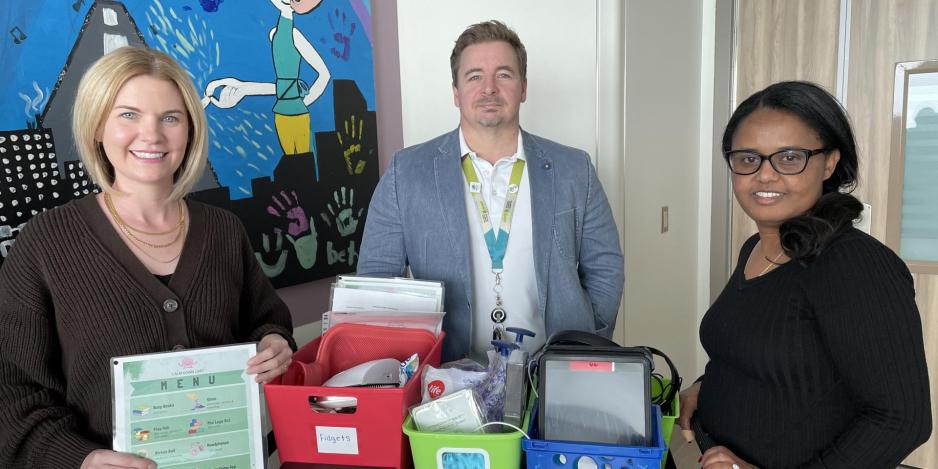 Three people pose for the camera with a calm down cart, a cart filled with activities to calm youth patients.