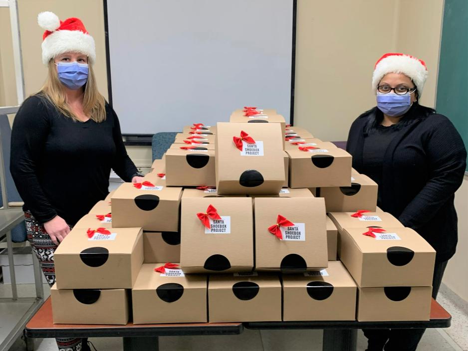 Two nurses wearing Santa hats and masks with a pile of boxes, each of which has text that reads “Santa Shoebox Project”. 