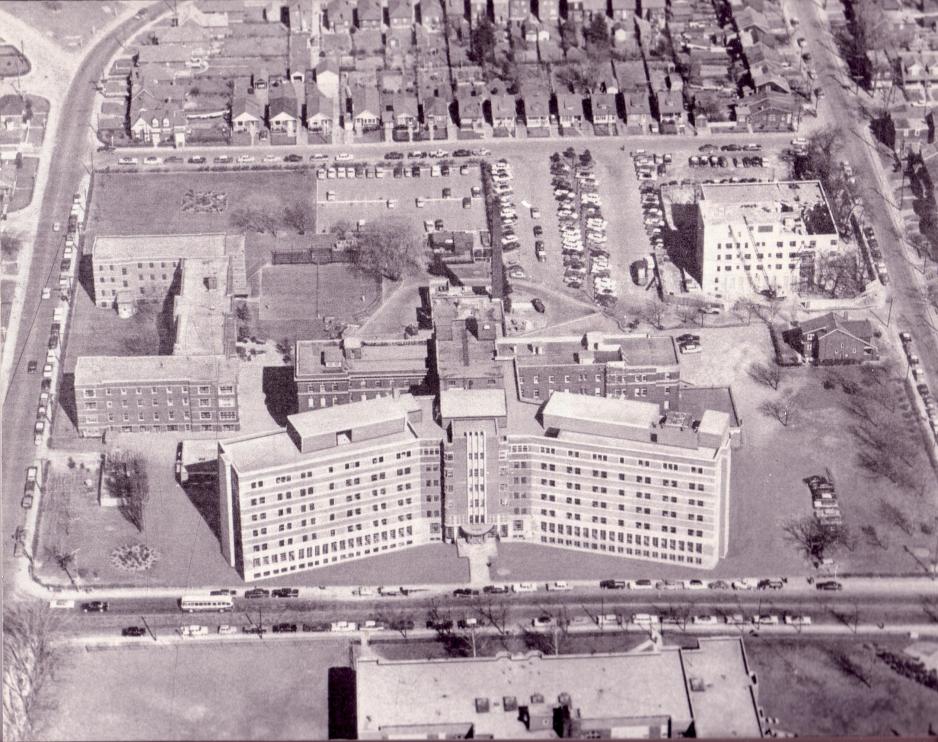 Black and white archival aerial photo of Michael Garron Hospital campus showing the curved wings from a central entrance, surrounded by buildings and residential neighbourhoods.