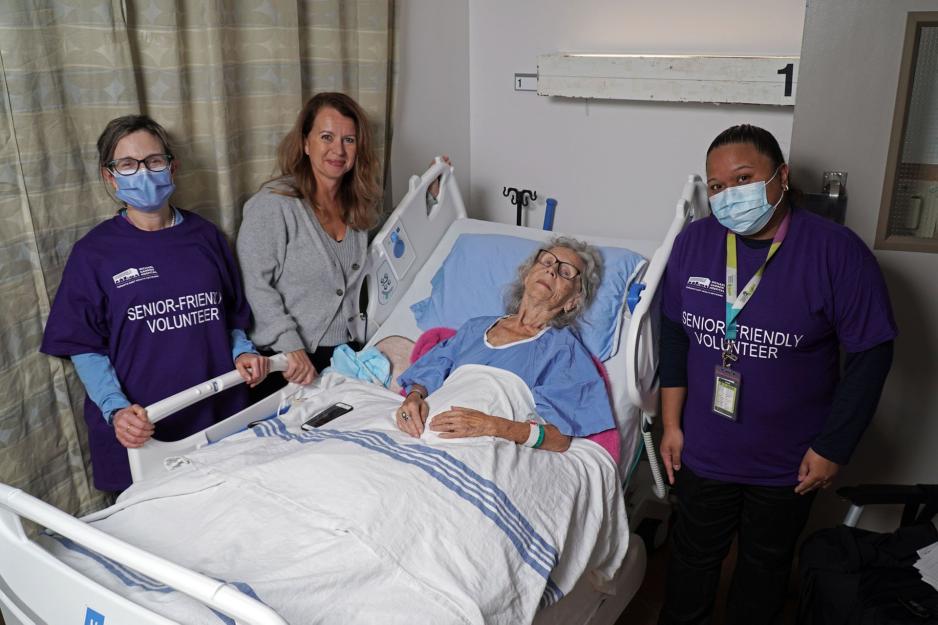 Senior friendly volunteers and hospital staff stand next to an elderly patient who is laying in a hospital bed